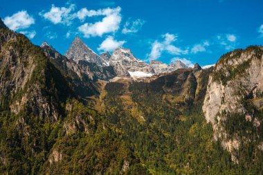 Çin, Balagezong 'da yemyeşil bir ormanı olan engebeli, karla kaplı bir dağ sırasının dramatik manzarası..