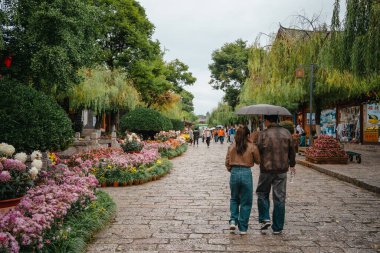 Lijiang, Çin - 23 Ekim 2024: Lijiang Eski Kasabası 'nda canlı çiçeklerle ve büyüleyici dükkanlarla çiçeklerle süslü yollarda romantik çiftler geziniyor.