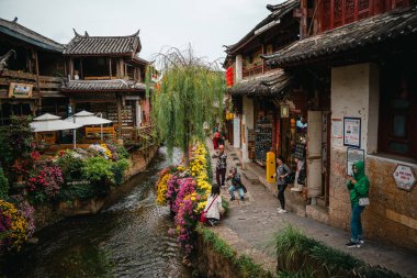 Lijiang, Çin - 23 Ekim 2024: Ziyaretçiler, geleneksel ahşap binalar ve söğüt ağaçları tarafından çerçevelenen Lijiang Eski Kasabası 'ndaki çiçekli bir kanalın yanında poz verip fotoğraf çekiyorlar..