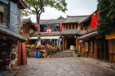 Lijiang Eski Kasabası 'nda sakin bir meydanda ahşap dükkanlar, saksı çiçekleri ve üzerinde bayraklar bulunan geleneksel Çin mimarisi yer alır..