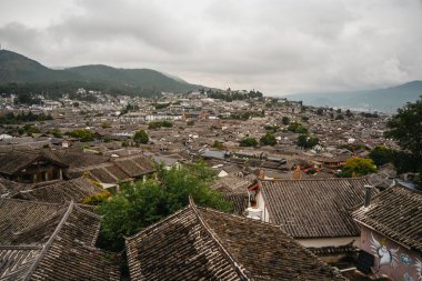 Lijiang Eski Kasabası 'nın geleneksel kiremitli çatılarına bakan arka planda dağlar ve bulutlu gökyüzü.