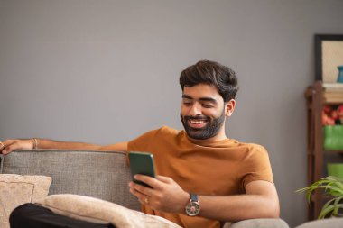 Genç adam oturma odasında koltukta dinlenirken cep telefonu kullanıyor.