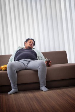 Şişman bir adam elinde patates kızartması ve kolayla odasında tembel tembel oturuyor..