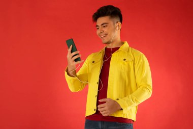Kırmızı arka planda cep telefonuyla müzik dinleyen mutlu bir gencin portresi