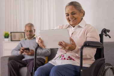 Yaşlı kadın tekerlekli sandalyede dijital tablet kullanıyor ve yaşlı adam da kanepede akıllı telefon kullanıyor.