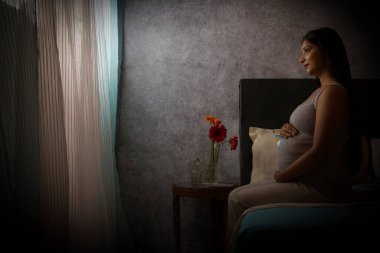 Hamile kadın evde yatakta otururken pencereden dışarı bakıyor.