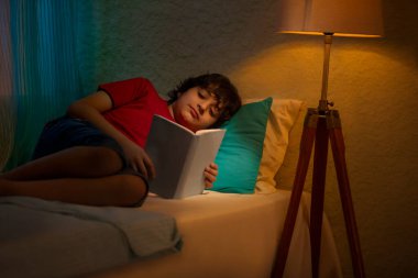 Küçük çocuk yatakta yatarken kitap okuyor.