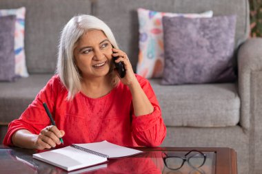 Old woman talking on mobile phone while writing on notepad at home clipart