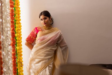 Beyaz sari giymiş Güney Hintli kadın duvara yaslanmış ve Onam vesilesiyle kameraya bakıyor.