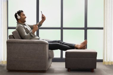 Genç işadamı kulaklıkla müzik dinliyor. Evdeki kanepede yatarken cep telefonu kullanıyor.