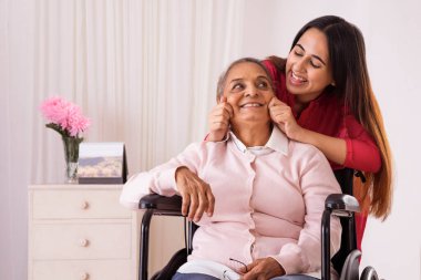Torunu, tekerlekli sandalyede oturan büyükannesinin yanağını çekiyor.