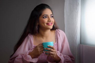 Pencerenin yanında oturan ve elinde bir fincan kahveyle dışarı bakan genç bir kadın.