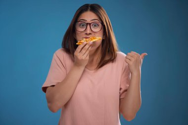 Gözleri kapalı bir kadın mavi arka planda bir dilim pizza yiyor.