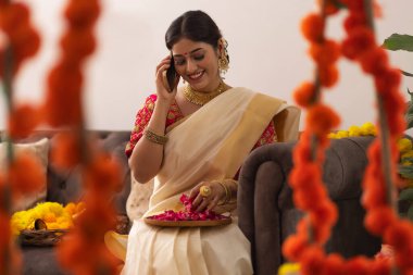 Geleneksel kıyafetli bir kadın elinde gül yapraklarıyla koltukta otururken cep telefonuyla konuşuyor.