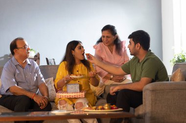 Kardeş, Raksha Bandhan 'da ailesiyle evde otururken ablasına laddoo yediriyor.