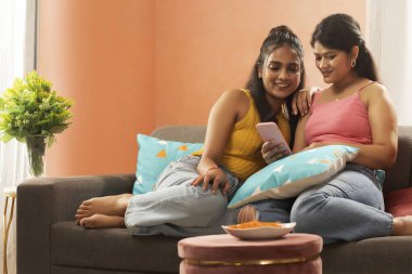 Oturma odasında kanepede dinlenirken cep telefonu kullanan iki gülümseyen kadın.