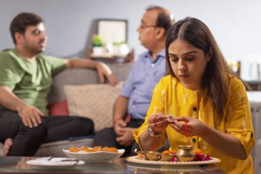 Raksha Bandhan vesilesiyle puja thali düzenleyen genç bir kadın. Babası ve abisi arkada oturuyor.
