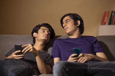 Oturma odasındaki kanepede uzanırken akıllı telefon kullanan iki gencin portresi.