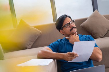 Orta yaşlı bir adam elinde bir kalem ve kağıt tutarak yere bakıyor.