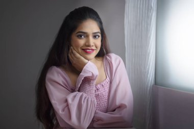 Genç bir kadın pencerenin yanında oturuyor ve elleri yanaklarında kameraya bakıyor.