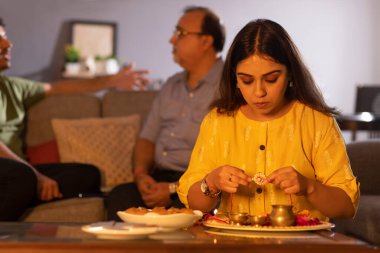 Young woman arranging puja thali on the occasion of Raksha Bandhan while her father and brother sitting behind clipart