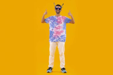 Gitar şeklinde gözlük takan ve baş parmaklarıyla el kol hareketi yapan komik bir genç adam.