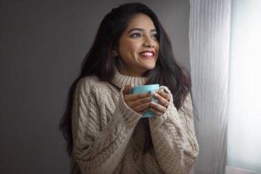 Kışlık kumaşlı genç bir kadın pencerenin yanında oturuyor ve elinde bir fincan kahveyle dışarıya bakıyor.