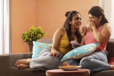 Oturma odasında kanepede dinlenirken cep telefonu kullanan iki gülümseyen kadın.