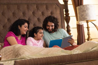 Happy family members watching photo album together in bedroom clipart