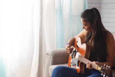 Oturma odasında kanepede oturan genç bir kadın gitar çalıyor.
