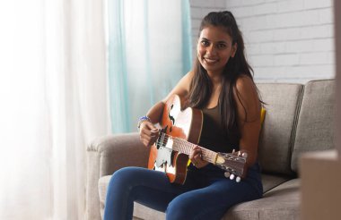 Oturma odasında kanepede oturan genç bir kadın gitar çalıyor.