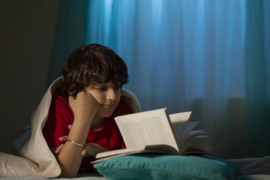 Çocuk yatakta battaniyeyle yatarken kitap okuyor.