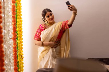 Beyaz sari giymiş Güney Hintli kadın, Onam olayında çiçek dekorasyonunun yanında dururken selfie çekiyor.