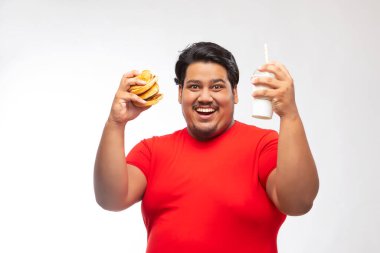 Portrait of a fat man holding a drink and burger in hand against plain background. clipart