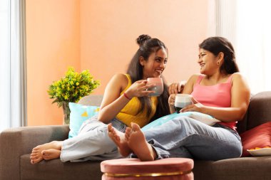 Oturma odasında kanepede dinlenirken kahve içen iki genç kadın.