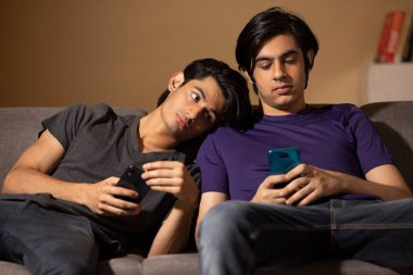 Oturma odasındaki kanepede uzanırken akıllı telefon kullanan iki gencin portresi.