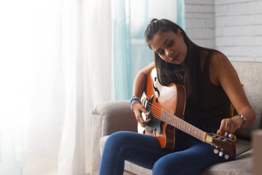 Oturma odasında kanepede oturan genç bir kadın gitar akort ediyor.