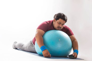 A fat man lying with closed eyes on a fitness ball kept on yoga mat with mobile against plain background. clipart