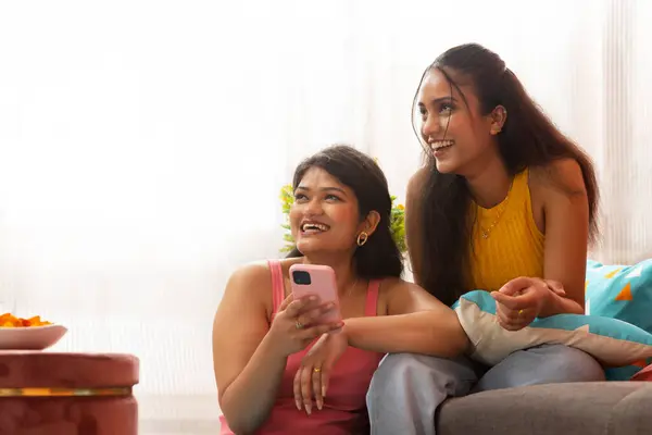 stock image Two smiling women using mobile phone together while sitting in living room