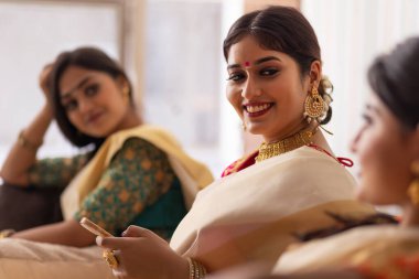 Geleneksel kıyafetli kadınlar koltukta birlikte otururken cep telefonu kullanır ve arkadaşlarıyla konuşurlar.