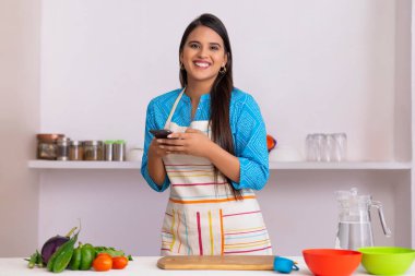 Beautiful Indian woman looking at camera with holding smartphone in kitchen clipart