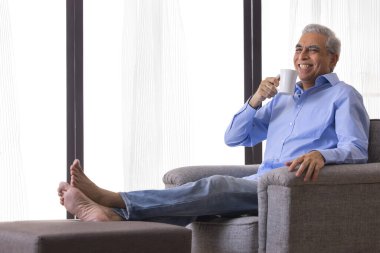 Evdeki kanepede dinlenirken kahve içen mutlu kıdemli adam.