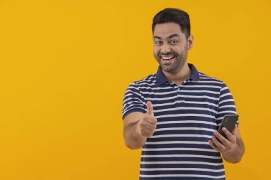 Elinde akıllı telefon olan genç bir adam kameranın önünde başparmağını gösteriyor.