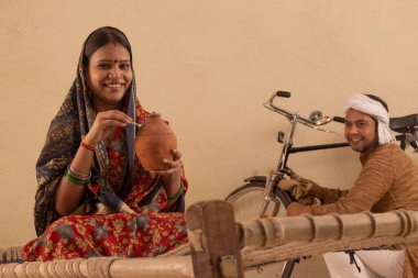 A RURAL WOMAN LOOKING AT CAMERA WHILE PUTTING MONEY IN PIGGY BANK WITH HUSBAND SITTING NEARBY clipart