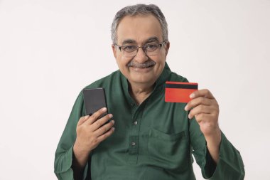 A HAPPY OLD MAN HOLDING MOBILE PHONE AND DEBIT CARD IN FRONT OF CAMERA clipart