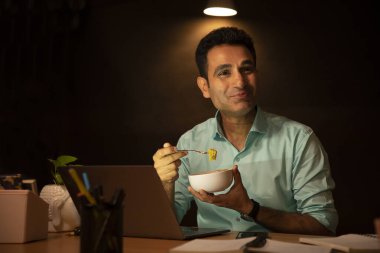 A CORPORATE PROFESSIONAL HAPPILY EATING MAGGI WHILE WORKING LATE AT NIGHT clipart