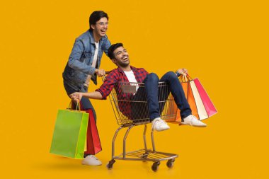 Two young boys laughing with one of them sitting in a trolley holding carrybags. clipart