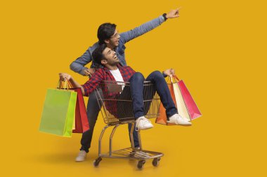 A young boy pointing elsewhere while moving a trolley with a boy holding carrybags sitting in it. clipart