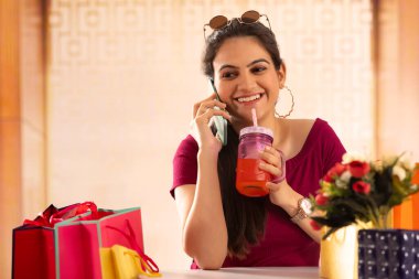 A girl with a drink in hand talking on phone after a shopping spree. clipart