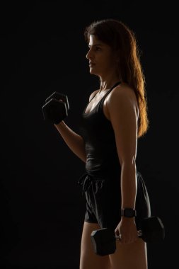 Koyu bir arkaplan önünde dambıllarla egzersiz yapan bir kadın..